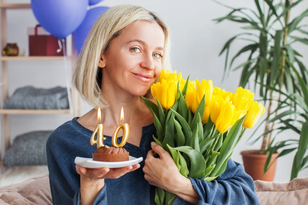 Smiling Middle aged woman with birthday cake and bouquet of flowers. Adult female celebration her birthday at home