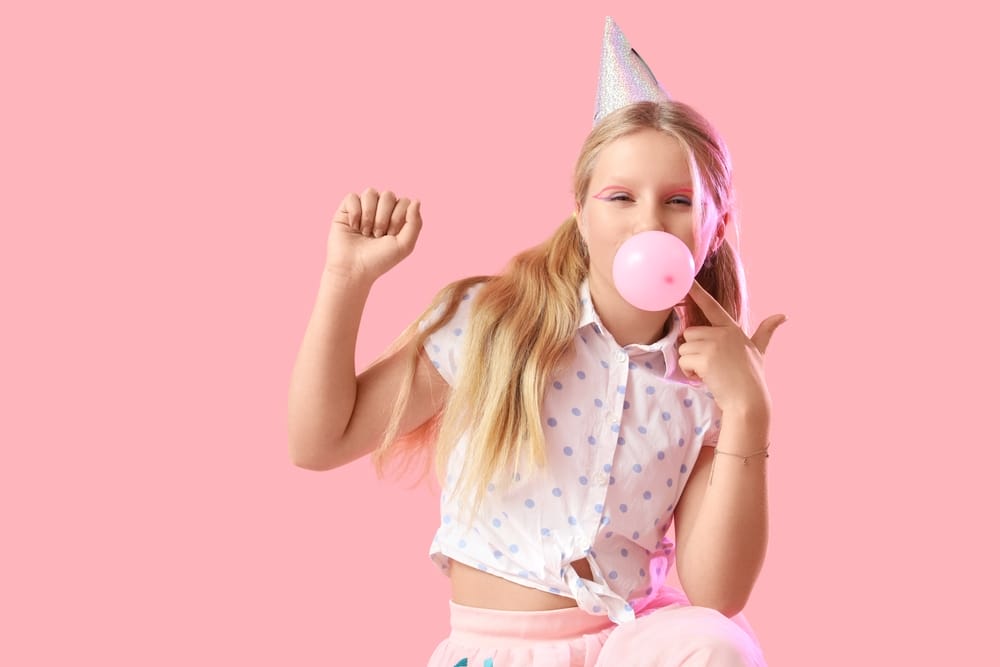 Cute trendy girl in birthday hat blowing bubble gum on pink background