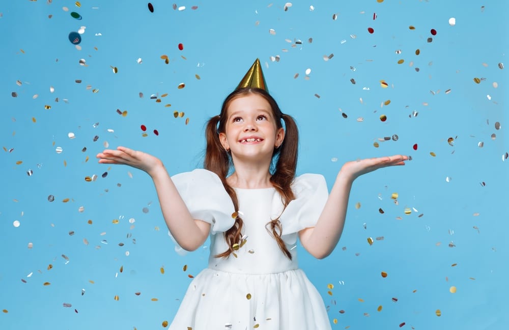 Young brunette woman in princess birthday hat, hands raised up and screaming, isolated on blue background, little girl
