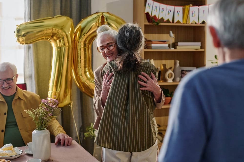Senior Caucasian woman smiling and hugging middle aged woman during birthday celebration with large gold 70 balloon in background, senior Caucasian man sitting at table nearby